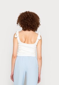 Top branco sem mangas com alças ajustáveis amarradas, detalhes franzidos na parte de trás, combinado com calças elásticas de cintura baixa em azul claro.