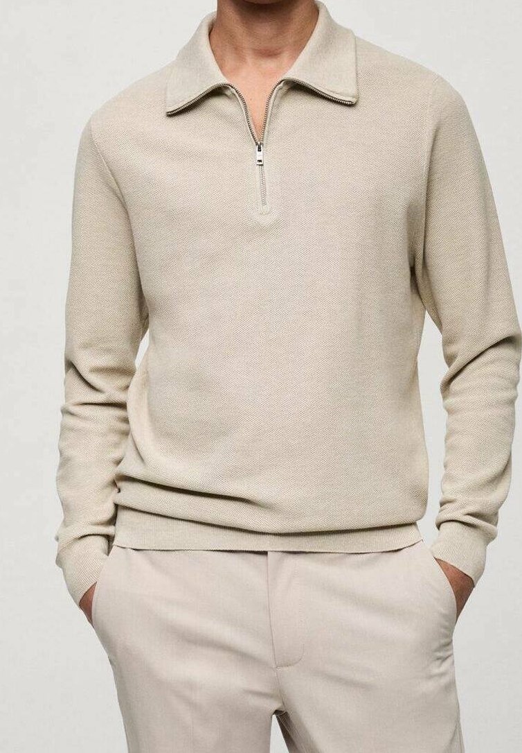 Homme portant un pull en tricot beige à manches longues avec col zippé et pantalon beige, mains dans les poches, devant un fond clair uni.