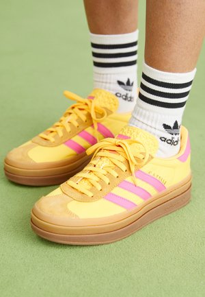 Benenden met witte Adidas-sokken met zwarte strepen en gele Adidas Gazelle sneakers met roze strepen op een groene vloer.