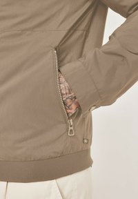 Khaki jacket with vertical ribbed texture, zippered pocket, fitted cuffs, and elastic hem. Visible tattoo on hand inside pocket.