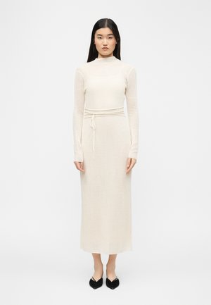Woman with long black hair wearing a fitted, ribbed cream-colored midi dress with long sleeves and black pointed-toe flats, standing on white background.