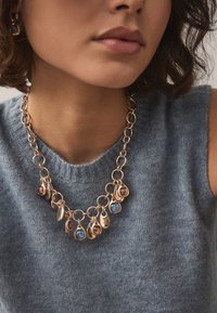 Gold-tone necklace with various shaped pendants, featuring blue and brown stones, worn over a light blue knitted sweater.