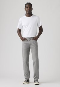 Άνδρας με πλεξουδάκια μαλλιά, στέκεται με τα χέρια στις τσέπες, φοράει λευκή μπλούζα, ανοιχτό γκρι τζιν και μαύρα αθλητικά παπούτσια μπροστά σε απλό φόντο.