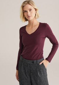 Blonde Frau trägt ein burgunderfarbenslangärmliges V-Ausschnitt-Oberteil und graue hoch taillierte Hose mit ihrer linken Hand in der Tasche vor einem beige Hintergrund.
