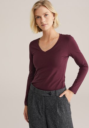 Blonde Frau trägt ein burgunderfarbenslangärmliges V-Ausschnitt-Oberteil und graue hoch taillierte Hose mit ihrer linken Hand in der Tasche vor einem beige Hintergrund.