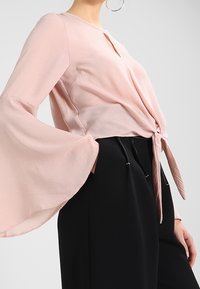 Vaaleanpunainen, pitkähihainen paita, jossa on kellotetut hihat ja vyötäröllä edessä solmittava nauha, yhdistettynä mustiin naruhousuihin. Sujuva rakenne.