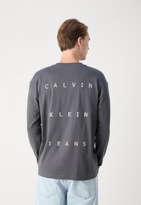Šedé bavlněné tričko s dlouhým rukávem a bílým blokovým písmem "CALVIN KLEIN JEANS" vytisknutým vertikálně na zadní straně. Volný střih.