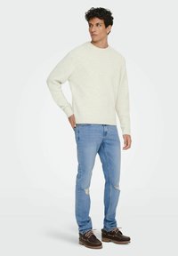 Suéter de punto crema claro con cuello redondo, combinado con jeans desgastados de color azul claro y zapatos oscuros de cuero con cordones y suela texturada.