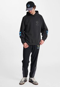 Svart hoodie med blå ränder på ärmarna, logotyp på bröstet. Matchande svarta träningsbyxor med sidodetaljer. Grå sneakers med blåa detaljer.