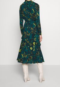 Langarmige jurk met een teal achtergrond, versierd met gele bloemmotieven en ruffles aan de zoom, gecombineerd met witte, chunky-heeled laarzen.