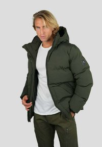 Homme portant une doudoune verte foncée avec capuche sur une chemise blanche et un pantalon vert assorti, sur fond gris uni.