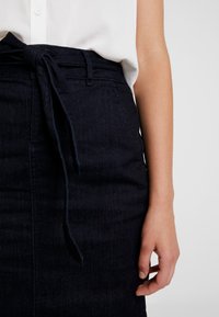 Jupe en denim bleu foncé avec une ceinture en tissu nouée, dotée d'une texture lisse et d'un design ajusté qui descend jusqu'au mi-mollet.