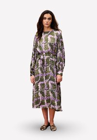 Vrouw met lang bruin haar, gekleed in een midi-jurk met lange mouwen met een abstract patroon in roze, groen en beige, en metallic ballerina's.