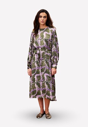 Femme aux longs cheveux bruns portant une robe midi à manches longues avec un motif abstrait rose, vert et beige, ainsi que des ballerines métalliques.