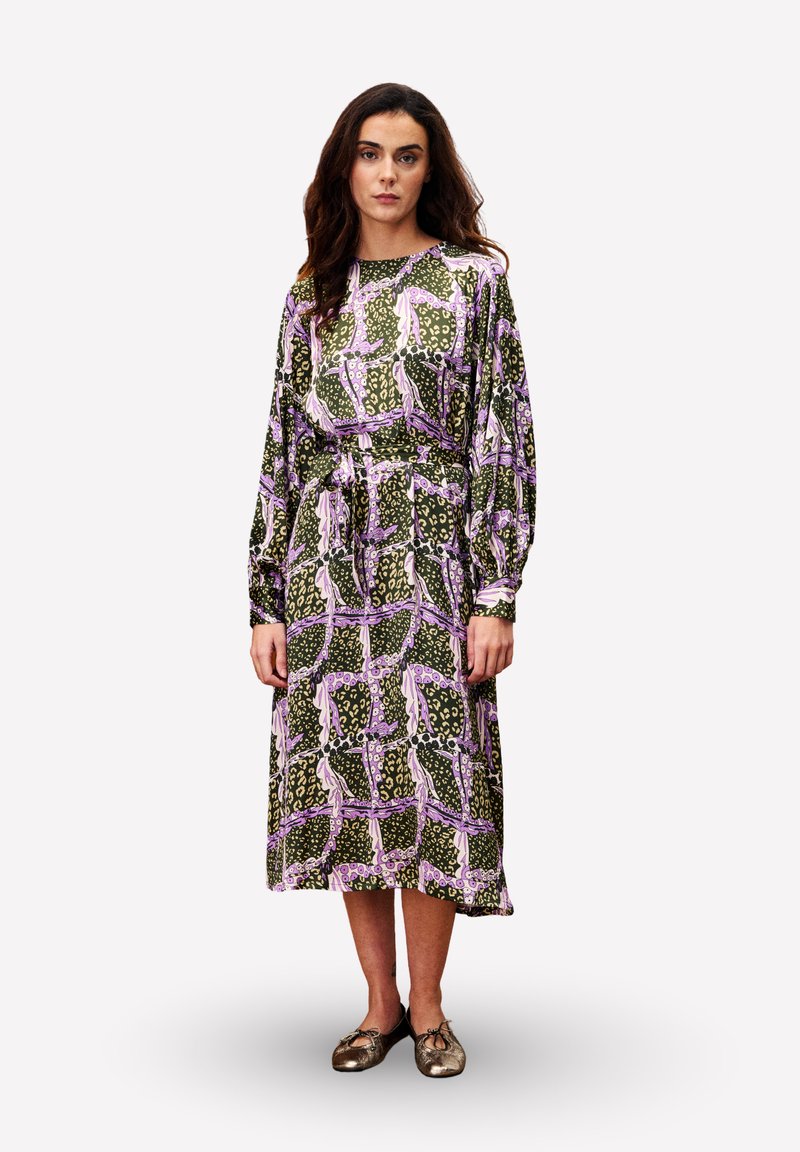 Vrouw met lang bruin haar, gekleed in een midi-jurk met lange mouwen met een abstract patroon in roze, groen en beige, en metallic ballerina's.