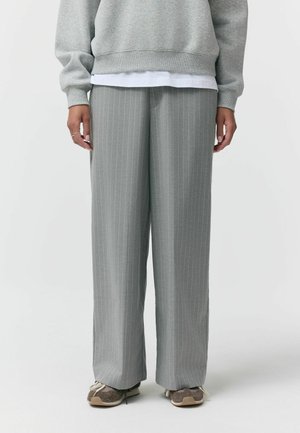 Osoba ubraną w szare spodnie z szerokimi nogawkami w cienkie paski, szarą bluzę na białą koszulę oraz brązowe trampki, stojąca.