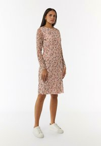 Blumenkleid in Pink, mit langen Ärmeln, Rundhalsausschnitt und knielangem Schnitt. Getragen mit weißen Sneakers, strukturierter Stoffdesign.