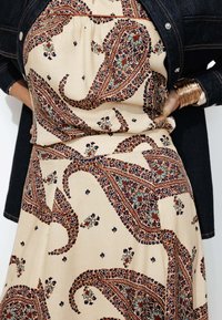 Robe beige avec un motif paisley en rouge, bleu et vert. Coupe ample, encolure froncée, accentuée par une veste en denim foncé.