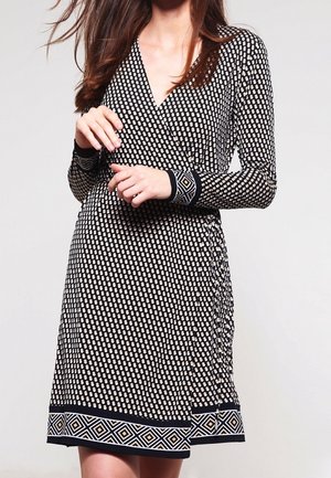 Femme portant une robe portefeuille noire et blanche à motifs, manches longues et bordure géométrique aux poignets et à l'ourlet, debout devant un fond uni.