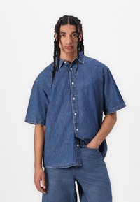 Cămașă din denim, mâneci scurte, culoare albastru deschis, închidere cu nasturi în față, guler ascuțit, croială lejeră, cusături vizibile, purtată cu pantaloni din denim asortați.