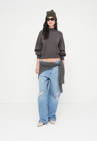 Πλεκτό πουλόβερ σε γκρι oversized, σκισμένο ανοιχτό μπλε τζιν με φαρδιά μπατζάκια, ζώνη από γκρι ύφασμα δεμένη, πράσινο σκουφάκι και μαύρα γυαλιά ηλίου.