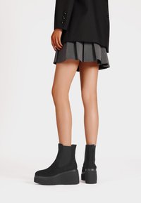 Stivaletti neri in suede con una suola chunky, pannelli elastici laterali e una finitura testurizzata, abbinati a una gonna grigia plissettata.