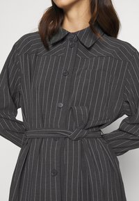 Vestido camisero gris oscuro con rayas verticales blancas. Presenta un cuello, cierre de botones, un bolsillo en el pecho y un cinturón a juego en la cintura.