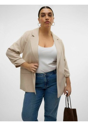 Nő bézs blézerben, fehér felsőben, kék farmerben, barna táskát tartva, egyszerű világos háttér előtt áll.