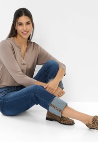Junge Frau mit dunklem Haar sitzt, trägt eine taupefarbene Bluse, hochgekrempelte blaue Jeans mit Leopardenmuster und passende Leopardenflats und lächelt.