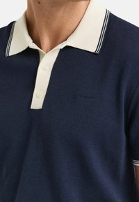 Námořnické polo tričko s kontrastním krémovým límcem a dvouknoflíkovou légou. Má tenké pruhy a vyšívané logo na hrudi.