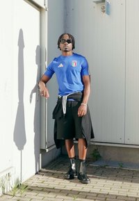 Μπλε αθλητική μπλούζα με το έμβλημα της Ιταλίας, μαύρο σορτς και μαύρα μποτάκια αστραγάλου· φοράει γυαλιά ηλίου και έχει δέσει το φούτερ στη μέση.