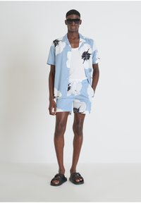 Camicetta azzurra a maniche corte con una grande stampa floreale bianca, abbinata a pantaloni corti coordinati e sandali neri, indossata sopra un top bianco.