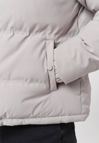Jachetă puf light gray, cu un buzunar lateral cu închidere pe bază de buton și manșete elasticizate, confecționată dintr-un material neted, ușor reflectorizant.