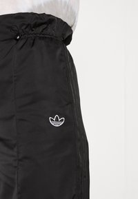 Svarta Adidas träningsbyxor med elastisk midja, tre svarta ränder nedtill på sidan och vitt trefoil-logga på låret mot en enfärgad bakgrund.