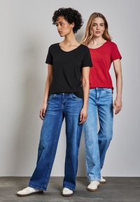 Dos mujeres llevan camisetas básicas; una en negro, la otra en rojo. Ambas tienen jeans relajados de tonos azul claro y oscuro, y zapatillas blancas casuales.