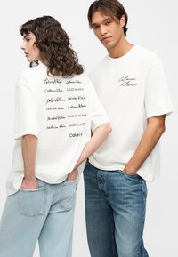 Δύο νέοι ενήλικες που φορούν λευκά t-shirts Calvin Klein και μπλε τζιν, στέκονται μπροστά σε ένα απλό φωτεινό φόντο.