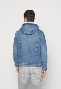 Blaues Jeansjackett mit Kapuze, ausgestattet mit einem weißen Fleece-Futter und verstellbaren Manschetten. Das Design umfasst Akzente an den Seitennähten und eine Rückenpasse.