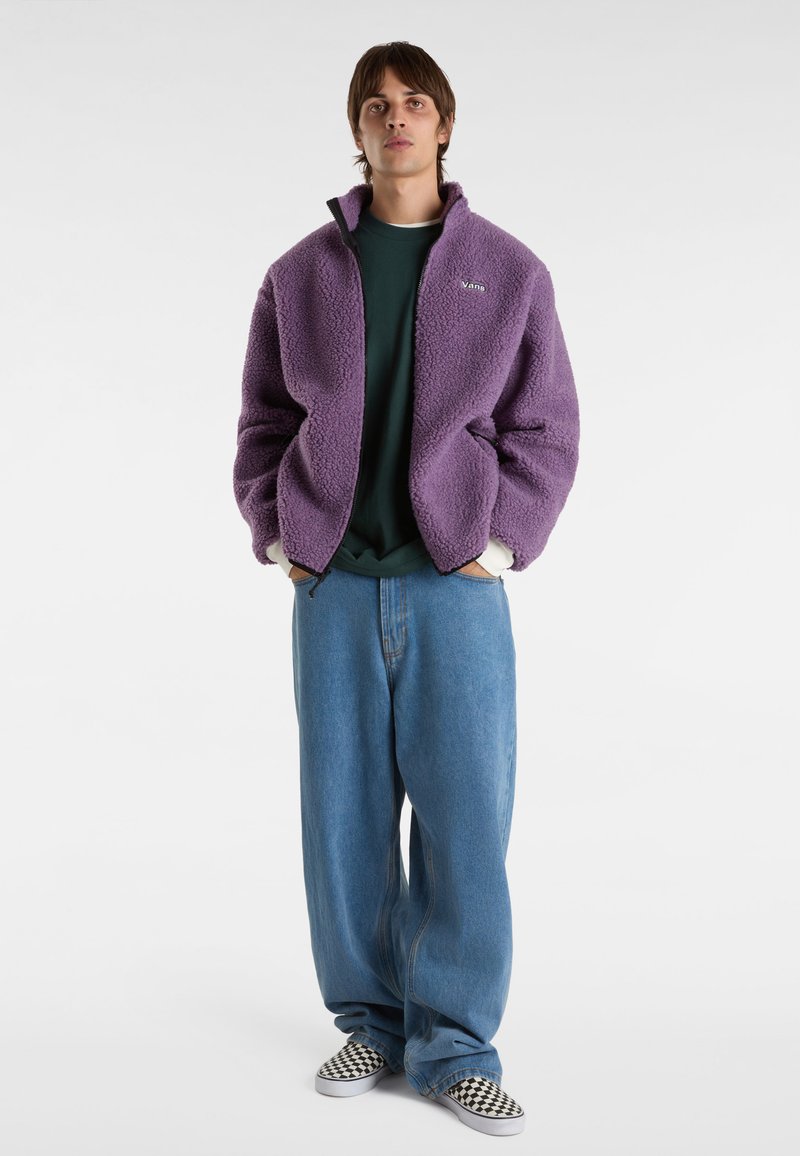 Lila Fleecejacke mit hohem Kragen, Reißverschluss vorne und Logo. Kombiniert mit einem grünen Hemd und lockeren blauen Jeans. Schwarz-weiße karierte Schuhe.