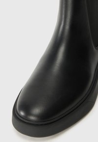 Botín de cuero negro con puntera redondeada, textura suave y panel lateral texturizado. Suela de goma con una ligera elevación.