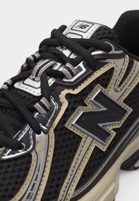 Gros plan sur une basket New Balance noire avec des touches beige et argent, des lacets noirs et à rayures, ainsi que des détails en tissu mesh.