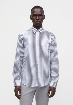 Uomo che indossa una camicia bianca a maniche lunghe con righe verticali nere e rosse e pantaloni grigio chiaro, in piedi contro uno sfondo chiaro e uniforme.