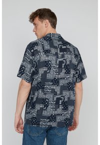 Rövid ujjú ing, sötétkék alapszínnel, fehér geometriai és paisley mintázattal, galléros dizájnnal és laza szabással. Hátulról látható.
