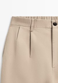 Pantaloni beige plissettati con una texture liscia, vita elasticizzata e un singolo bottone nero in alto. Design su misura con tasche laterali.