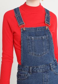 Salopette di jeans blu con componenti in metallo, spalline regolabili e tasca frontale, abbinata a un maglione aderente a collo alto rosso.