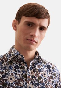 Camicia bianca a bottoni con un mix di fiori blu, verdi e marroni su uno sfondo chiaro. Con colletto e tessuto testurizzato.