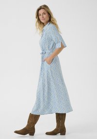 Blommig midi-klänning i ljusblått tyg med gula och vita blommor, bältad vid midjan och med sidofickor. Matchad med bruna mockakängor.