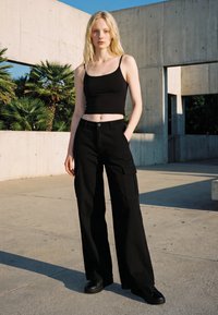 Ung kvinde med langt blondt hår iført en sort crop top og vide sorte cargobukser står udendørs nær betontænder.