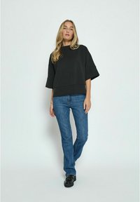Black oversized sweatshirt with wide sleeves, paired with blue jeans. Model stands in a neutral background, showcasing the outfit's fit and design.