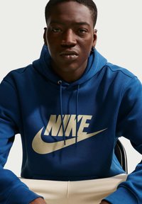 Junger Mann in blauem Nike-Hoodie mit beigem Logo, sitzt mit Händen auf den Knien vor einfachem, hellem Hintergrund.