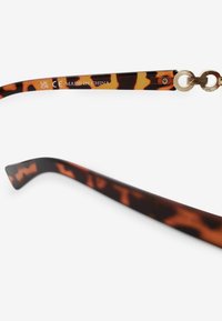 Gafas de carey con un patrón en marrón oscuro y naranja. Presenta puntas curvadas y un acento de hardware dorado en la bisagra. Texto en el brazo visible.